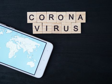 Coronavirus word made of wood block and world map on smartphone screen. Coronavirus text on dramatic atmosphere black wooden table. Coronavirus concept top view, copy space