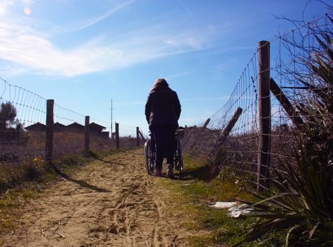 lonely person from behind who pushes a wheelchair alone along a sandy path that leads to the sea in the dunes in tuscany