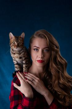 Fashion photo of pretty girl with bengal kitten on shoulder. Bengal cat with beautiful woman on  blue background. Girl and kitten seductively looks at the viewer.