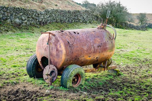 A rusty, corroded slurry tanker with punctured tyres sits abandoned in a muddy field in the Irish countryside.