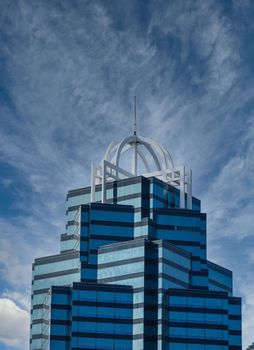 A glass office tower of light and dark blue with white trim on top under partly cloudy skies