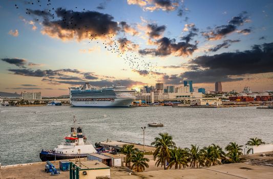 A busy tropical harbor with a large luxury cruise ship docked along with a tug boat and smaller boats