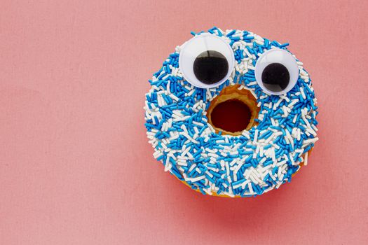 Vanilla Dip Donut with Black Wiggle Googly Eyeballs on a Coloured blue and white Sprinkles on a pink background.