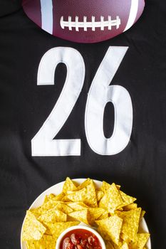 An American football with organic nacho chips and mild salsa on a white black football jersey with the 26 number on vertical view