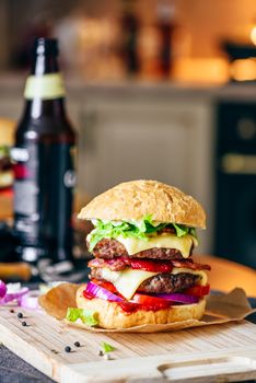 Cheeseburger with Two Beef Patties, Cheddar Cheese, Bacon, Iceberg Lettuce, Sliced Tomatoes and Red Onion. Bottle of Beer and Some Ingredients on Table.