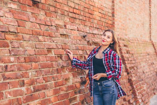 Portrait of young woman in checkered shirt and blue jeans standing against brick wall