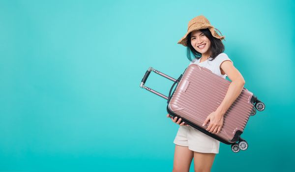 Portrait happy Asian beautiful young woman standing wear white t-shirt, holidays travel concept, her hugging suitcase bag and looking to camera, shoot photo in a studio on a blue background