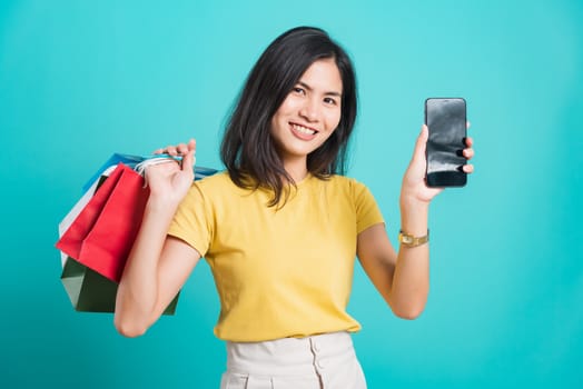 Portrait happy Asian beautiful young woman smile white teeth standing wear yellow t-shirt, She holding shopping bags and using a mobile phone, studio shot on blue background with copy space for text