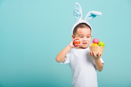 Asian cute little child boy smile beaming wearing bunny ears and a white T-shirt, standing to hold a basket with full Easter eggs. And other hand holds an easter egg on blue background with copy space