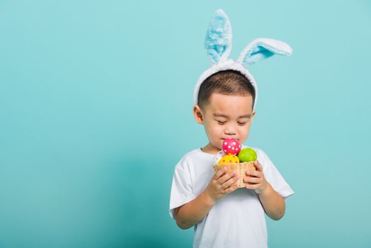 Asian cute little child boy smile beaming wearing bunny ears and a white T-shirt, standing to hold a basket with full Easter eggs on blue background with copy space