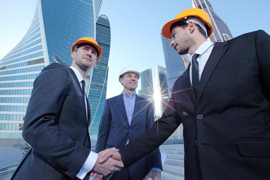 Team of successful young engineers and business partners shaking hands during a meeting on a construction site over skyscrapers background. Architecture and teamwork concept.