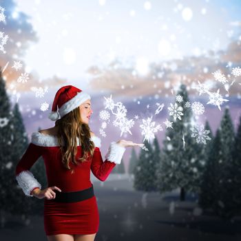 Pretty girl in santa costume holding hand out against snowy landscape with fir trees