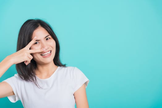 Portrait Asian beautiful happy young woman smile white teeth wear white t-shirt standing showing two finger making v-sign near the eye, on a blue background with copy space
