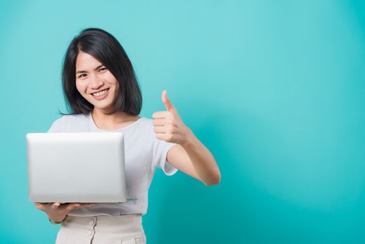 Asian beautiful young woman smile white teeth standing to holding laptop computer and showing thumb up while shoot photo in studio on blue background with copy space for text