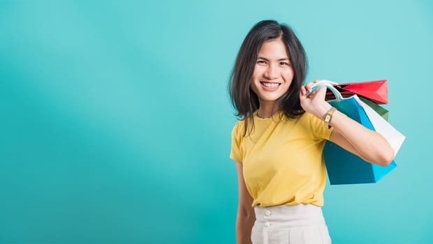 Portrait happy Asian beautiful young woman smile white teeth standing wear yellow t-shirt, She holding shopping bags on hand her looking camera, shoot photo in studio on blue background with copyspace