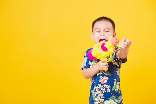 Portrait happy Asian cute little children boy smile standing so happy wearing flower shirt in Songkran festival day holding water gun, studio shot on yellow background with copy space