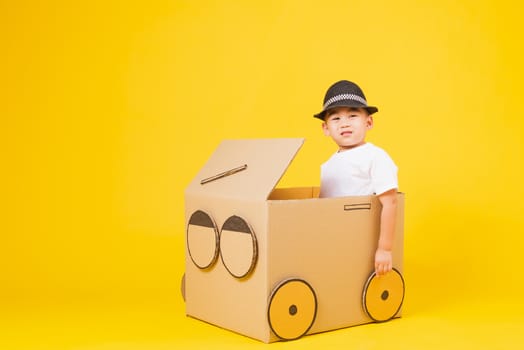 Portrait happy Asian cute little children boy smile so happy wearing white T-shirt driving car creative by cardboard, studio shot on yellow background with copy space