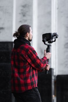 Young Professional videographer holding professional camera on 3-axis gimbal stabilizer. Pro equipment helps to make high quality video without shaking. Cameraman wearing red shirt making a videos