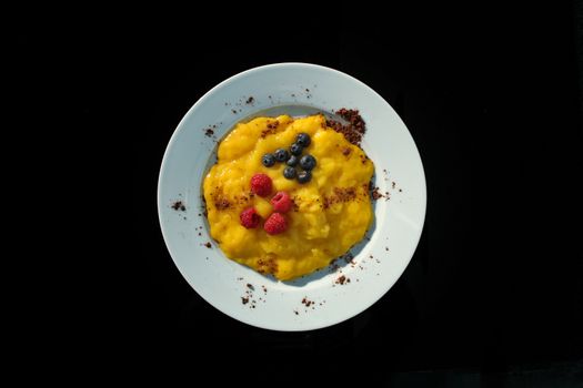 Yellow pudding with blueberry and raspberries with chocolate chips on a white plate and black background