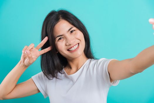 Portrait happy Asian beautiful young woman smile white teeth standing wear white t-shirt, She makes selfie front camera smartphone showing v-sign, looking to camera studio shot on blue background