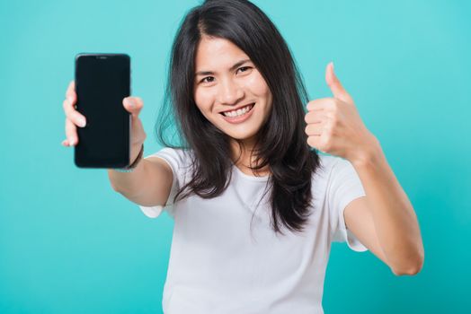 Portrait Asian beautiful happy young woman standing smile, holding blank screen mobile phone and showing thumbs up gesture, shoot photo in studio focus face on blue background, with copy space