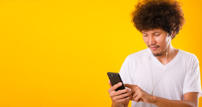 Happy asian handsome man with curly hair he smiling enjoying listening to music on earphones using a mobile smartphone isolate on yellow background