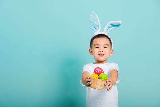 Asian cute little child boy smile beaming wearing bunny ears and a white T-shirt, standing to hold a basket with full Easter eggs on blue background with copy space