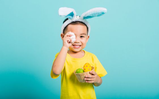 Asian cute little child boy smile beaming wearing bunny ears and a yellow T-shirt, standing to holds colored easter eggs instead of eyes on blue background with copy space for text