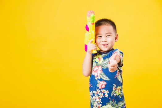 Portrait happy Asian cute little children boy smile standing so happy wearing flower shirt in Songkran festival day holding water gun, studio shot on yellow background with copy space