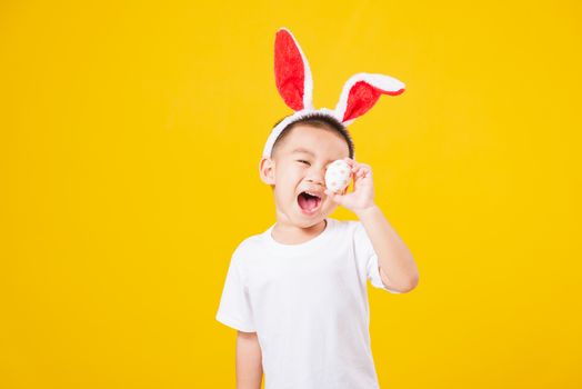 Portrait happy Asian cute little children boy smile standing so happy wearing white T-shirt and bunny ears in Easter festival day holding easter eggs, studio shot on yellow background with copy space