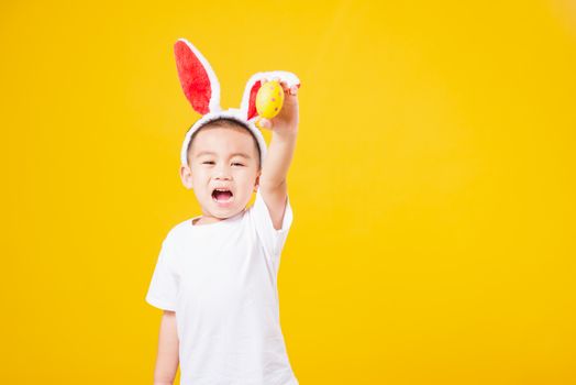 Portrait happy Asian cute little children boy smile standing so happy wearing white T-shirt and bunny ears in Easter festival day holding easter eggs, studio shot on yellow background with copy space