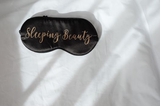 Silk black sleep mask with inscription Sleeping beauty on white rumpled sheets. Top view, flat lay. Horizontal. Copy spase. Concept of rest, awakening, sleep. For social media, blog. Minimal style.