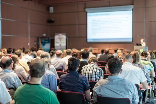 Female speaker giving presentation in lecture hall at university workshop. Audience in conference hall. Rear view of unrecognized participant in audience. Scientific conference event.