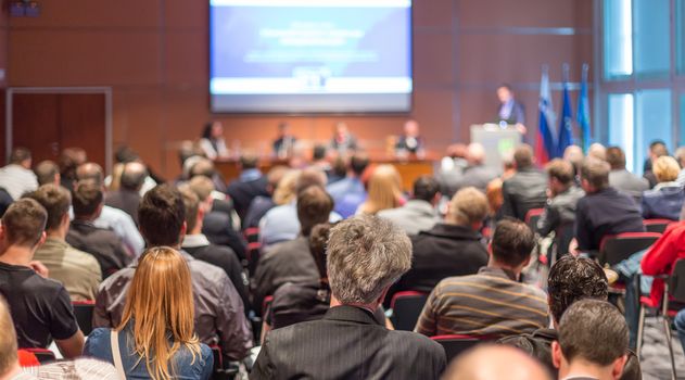 Business and entrepreneurship symposium. Speaker giving a talk at business meeting. Audience in conference hall. Rear view of unrecognized participant in audience.