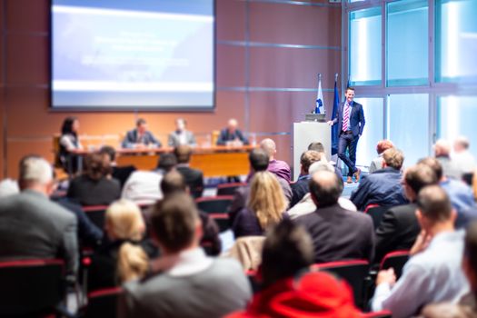 Business and entrepreneurship symposium. Speaker giving a talk at business meeting. Audience in conference hall. Rear view of unrecognized participant in audience.