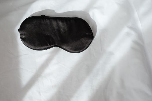 Silk black sleep mask without inscription on white rumpled sheets. Top view, flat lay. Horizontal. Copy spase. Concept of rest, awakening, sleep. For social media, blog. Minimal style.