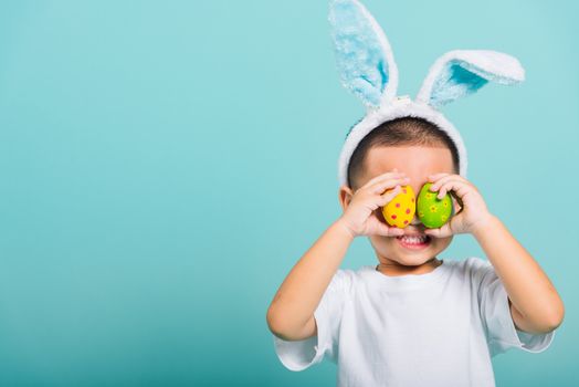 Asian cute little child boy smile beaming wearing bunny ears and a white T-shirt, standing to holds colored easter eggs instead of eyes on blue background with copy space for text