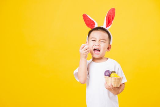 Portrait happy Asian cute little children boy smile standing so happy wearing white T-shirt and bunny ears in Easter festival day holding easter eggs, studio shot on yellow background with copy space