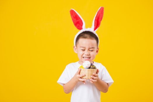 Portrait happy Asian cute little children boy smile standing so happy wearing white T-shirt and bunny ears in Easter festival day holding easter eggs, studio shot on yellow background with copy space