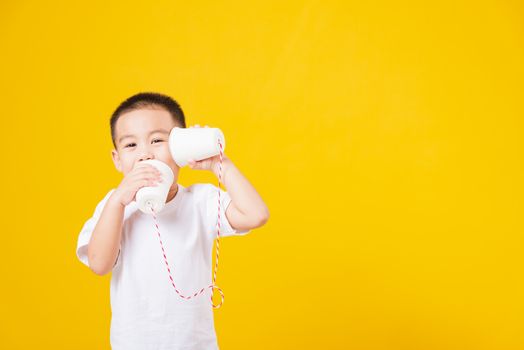 Portrait happy Asian cute little children boy smile standing so happy wearing white T-shirt playing paper can telephone, studio shot on yellow background with copy space
