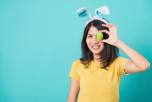 Portrait Asian beautiful happy young woman smile white teeth wear yellow t-shirt standing with bunny ears and holding Easter eggs near the eye, on blue background with copy space