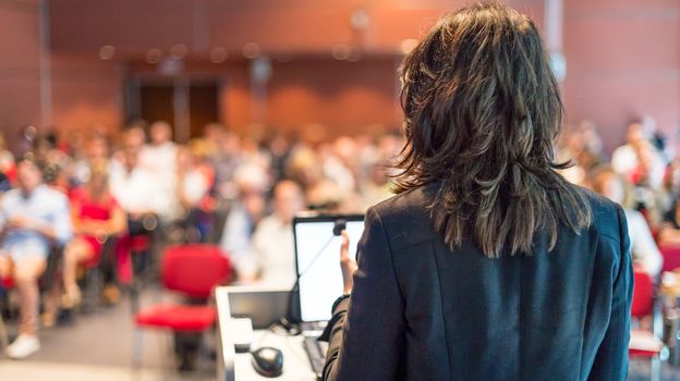 Female speaker giving a talk on corporate business conference. Unrecognizable people in audience at conference hall. Business and Entrepreneurship event.