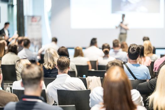 Business and entrepreneurship symposium. Speaker giving a talk at business meeting. Audience in conference hall. Rear view of unrecognized participant in audience.