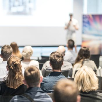 Business and entrepreneurship symposium. Speaker giving a talk at business meeting. Audience in conference hall. Rear view of unrecognized participant in audience.