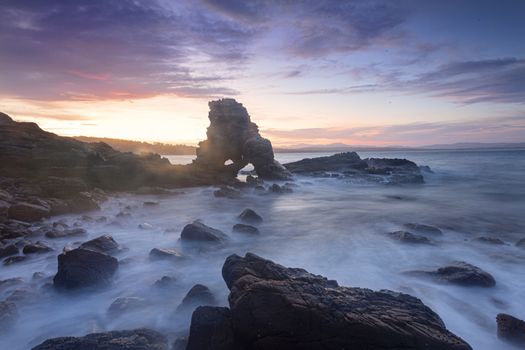 Sun setting behind the arched cave rock, its rays highlighting the sea spray carried on the wind as some bigger than usual waves washed in to this little stony cove giving a magical mystical scene