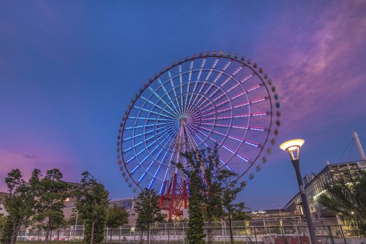 Odaiba colorful tall Palette Town Ferris wheel named Daikanransha visible from the central urban area of Tokyo in the summer sunset purple blue sky. Passengers can see the Tokyo Tower, the twin-deck Rainbow Bridge, and Haneda Airport, as well as central Tokyo during their ride.