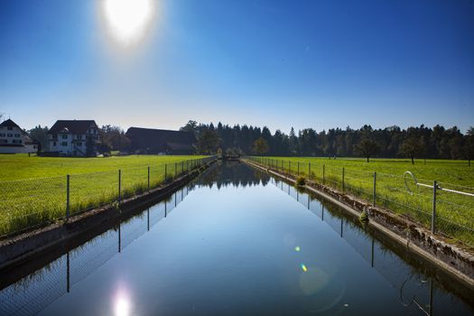 calm river in green countryside in Switzerland, with a fence with sun and sun reflection in water