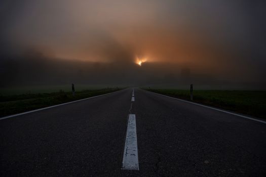 road towards horizon during sunrise with fog. Showing white middle line and dark hill