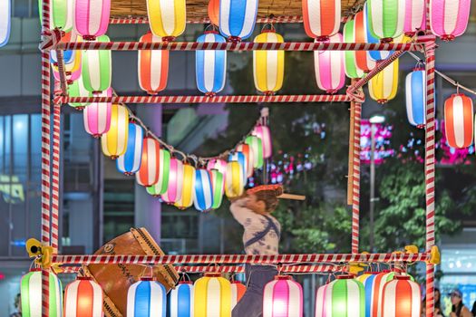 View of the square in front of the Nippori train station decorated for the Obon festival with a yagura tower illuminated with paper lanterns where a girl in traditional costume is playing taiko drum in the summer night of Arakawa district in Tokyo.