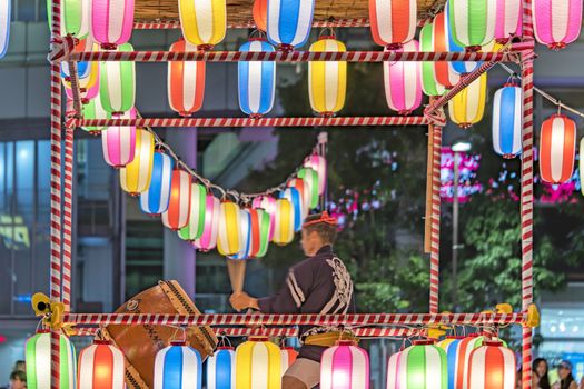 View of the square in front of the Nippori train station decorated for the Obon festival with a yagura tower illuminated with paper lanterns where a man in traditional costume is playing taiko drum in the summer night of Arakawa district in Tokyo.